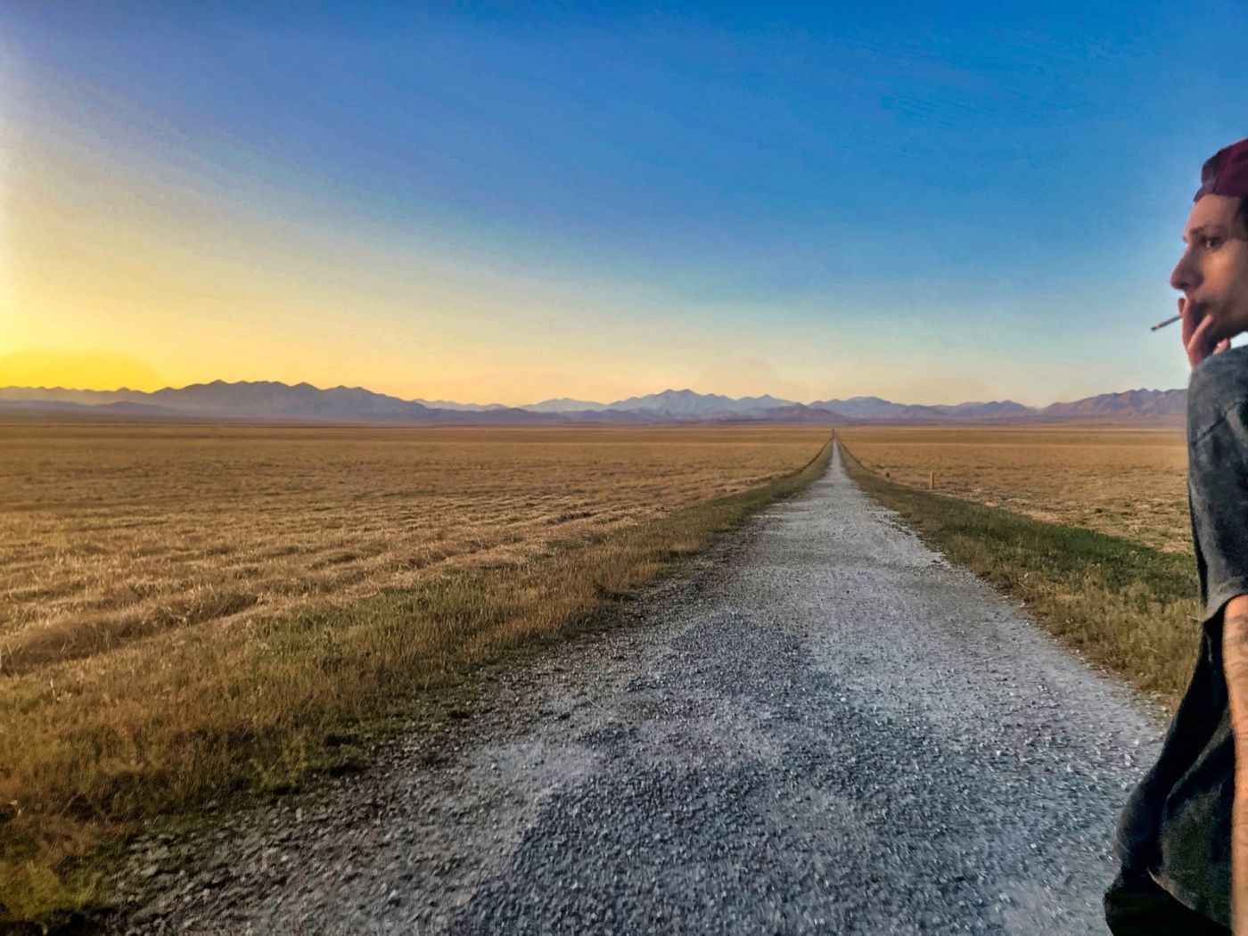 An endless gravel road through golden fields toward the mountains at sunset